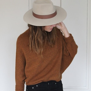 以下が含まれることがあります： 茶色のセーター、黒いジーンズ、茶色のバンドが付いたベージュのフェドーラ帽を着用した女性