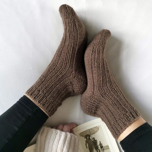 Pode incluir: Um par de meias de l&atilde; castanhas com um padr&atilde;o de nervuras. As meias s&atilde;o usadas por uma pessoa sentada em uma superf&iacute;cie branca com um livro no colo. A capa do livro mostra uma foto em preto e branco de uma mulher segurando um guarda-chuva em uma praia.