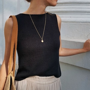 Könnte beinhalten: Eine Frau trägt ein schwarzes ärmelloses Strickoberteil mit einer goldenen Kettenkette und einem runden Anhänger. Sie trägt auch eine beige Hose und eine braune Ledertasche.