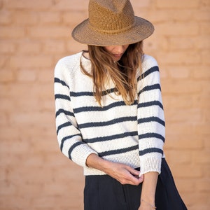 以下が含まれることがあります： 白と黒のストライプのセーターを着て、茶色のつば広帽子をかぶった女性。黒いスカートを履いていて、長い茶色の髪をしている。