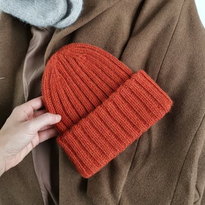 Könnte beinhalten: Eine braune Strickmütze mit gerippter Struktur. Die Mütze liegt auf einem braunen Mantel.