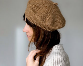 May include: A tan-colored knitted beret. The beret is made of a textured yarn and has a classic, round shape. The beret is worn with a light gray sweater and a leopard print scrunchie.