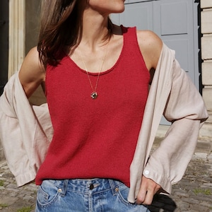 Könnte beinhalten: Eine Frau trägt ein rotes Tanktop, blaue Jeans und eine Sonnenbrille. Ein helles Hemd ist über die Schultern gelegt. Eine goldene Halskette mit Anhänger ist sichtbar. Im Hintergrund ein Steingebäude mit grauer Tür.