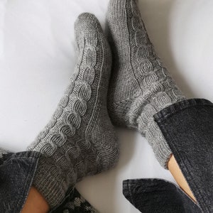 Puede incluir: Un par de calcetines de punto grises con un patrón de trenza. Los calcetines se llevan puestos en los pies de una persona y están parcialmente metidos en unos vaqueros negros.