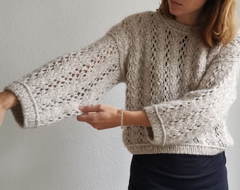 May include: A beige knitted sweater with a delicate openwork pattern. The sweater has a relaxed fit and long sleeves.