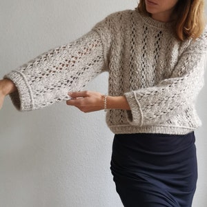 Puede incluir: Un suéter de punto beige con un delicado patrón de calado. El suéter tiene un corte holgado y mangas largas.