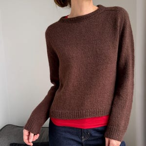 Peut inclure: Un pull marron à col rond et manches longues. Le pull en tricot a un ourlet et des poignets côtelés. Un t-shirt rouge est porté en dessous, associé à un jean bleu.