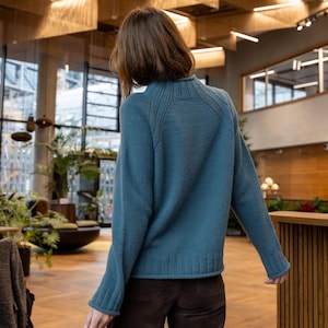 Könnte beinhalten: Ein teal-farbener Pullover mit geripptem Saum und Bündchen. Der Pullover hat einen Rundhalsausschnitt und lange Ärmel. Die Person auf dem Bild trägt eine braune Cordhose. Der Hintergrund ist ein modernes Interieur mit Holzakzenten und großen Fenstern.