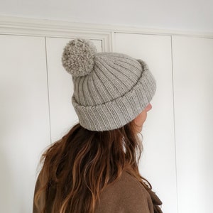 Könnte beinhalten: Eine graue Strickmütze mit einem flauschigen Bommel oben. Die Mütze wird von einer Person mit langen braunen Haaren getragen.
