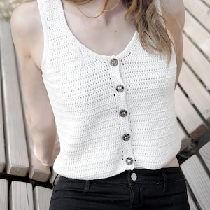 以下が含まれることがあります： ボタン留めのフロントを持つ白いクロシェタンクトップ。タンクトップは、長い茶色の髪と茶色の麦わら帽子をかぶった人が着用しています。