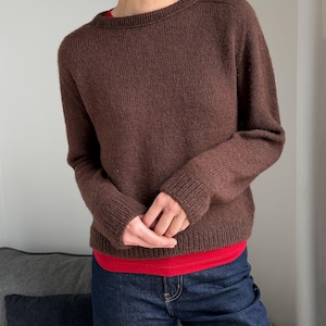Puede incluir: Un suéter de punto de color marrón oscuro de manga larga con cuello redondo y puños y dobladillo acanalados. El suéter se lleva sobre una camiseta interior de color rojo brillante y se combina con vaqueros de mezclilla azul oscuro.