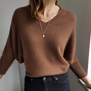 以下が含まれることがあります： Vネックと長袖のブラウンニットセーター。セーターはブラックジーンズと、丸いペンダントの付いたゴールドネックレスと合わせています。