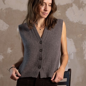 以下が含まれることがあります： Vネックとボタン留めのグレーのノースリーブニットベスト。 ベストは柔らかい素材で作られています。 モデルは茶色のコーデュロイパンツとベストを着用しています。