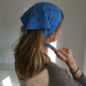 May include: A sky blue knitted bandana featuring a diamond openwork design. The bandana is tied at the back of the head. The person is wearing a brown sweater. The ends of the ties are visible.
