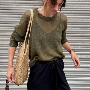 Könnte beinhalten: Ein olivgrüner Langarmpullover mit grob gestrickter Textur und Rundhalsausschnitt, kombiniert mit einer schwarzen Hose mit Kordelzug und einer beigen, geflochtenen Strohtasche. Der Pullover hat eine halbtransparente Struktur mit Rippkragen.