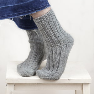 Pode incluir: Um par de meias de l&atilde; cinzas com textura canelada. As meias s&atilde;o usadas nos p&eacute;s de uma pessoa e est&atilde;o apoiadas em um banquinho de madeira branco.