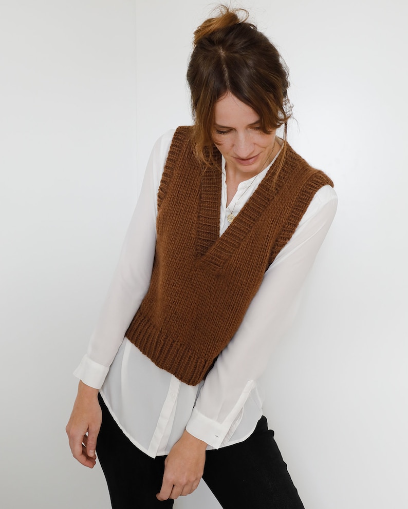 May include: A brown knitted vest with a V-neckline. The vest is worn over a white button-down shirt.