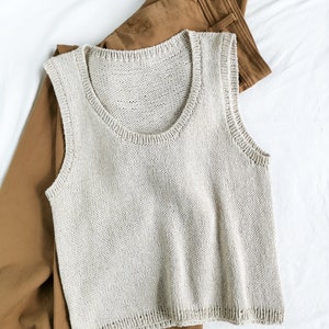 Pode incluir: Um top de malha bege claro sem mangas com decote redondo. O top está sobre calças marrons.