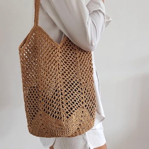 Peut inclure: Un sac fourre-tout tissé, de couleur beige naturel. Le sac a une forme rectangulaire avec une large ouverture et une longue sangle pour le porter à l'épaule. Le design du sac présente un motif géométrique en bas.