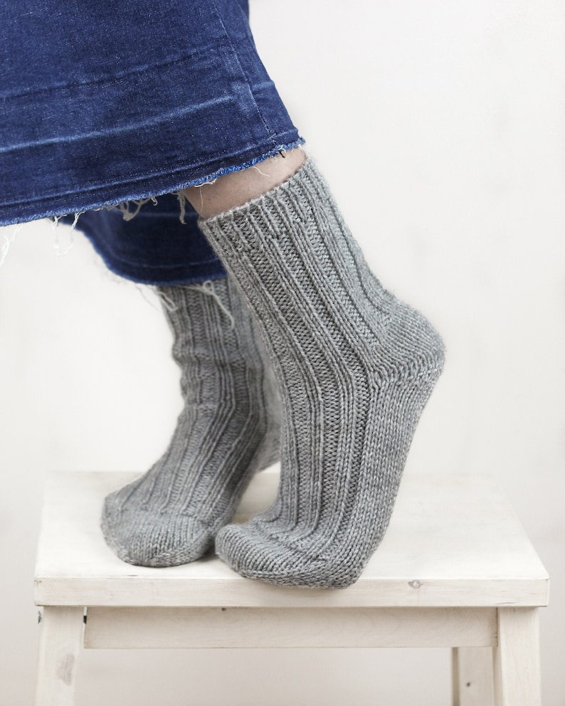 Pode incluir: Um par de meias de l&atilde; cinzas com textura canelada. As meias s&atilde;o usadas nos p&eacute;s de uma pessoa e est&atilde;o apoiadas em um banquinho de madeira branco.