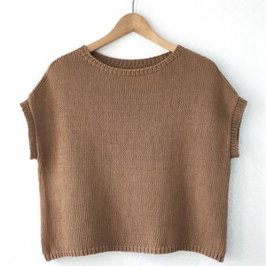 Pu&ograve; includere: Un top di maglia marrone senza maniche con scollo rotondo, appeso a un appendiabiti di legno su uno sfondo bianco.