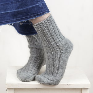 May include: A pair of gray knitted socks with a ribbed texture. The socks are worn on a person's feet and are resting on a white stool.