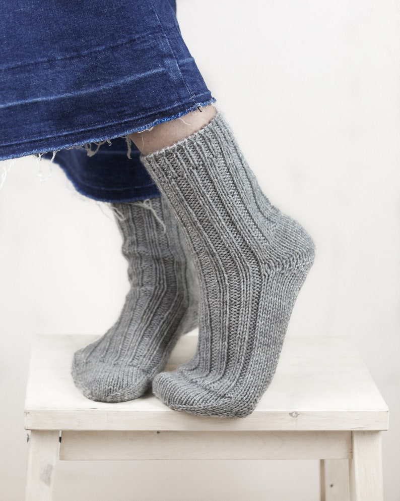 May include: A pair of gray knitted socks with a ribbed texture. The socks are worn on a person's feet and are resting on a white stool.