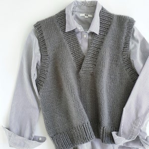 May include: A gray knitted vest with a V-neck and a ribbed texture. The vest is hanging on a hanger and is layered over a striped shirt.