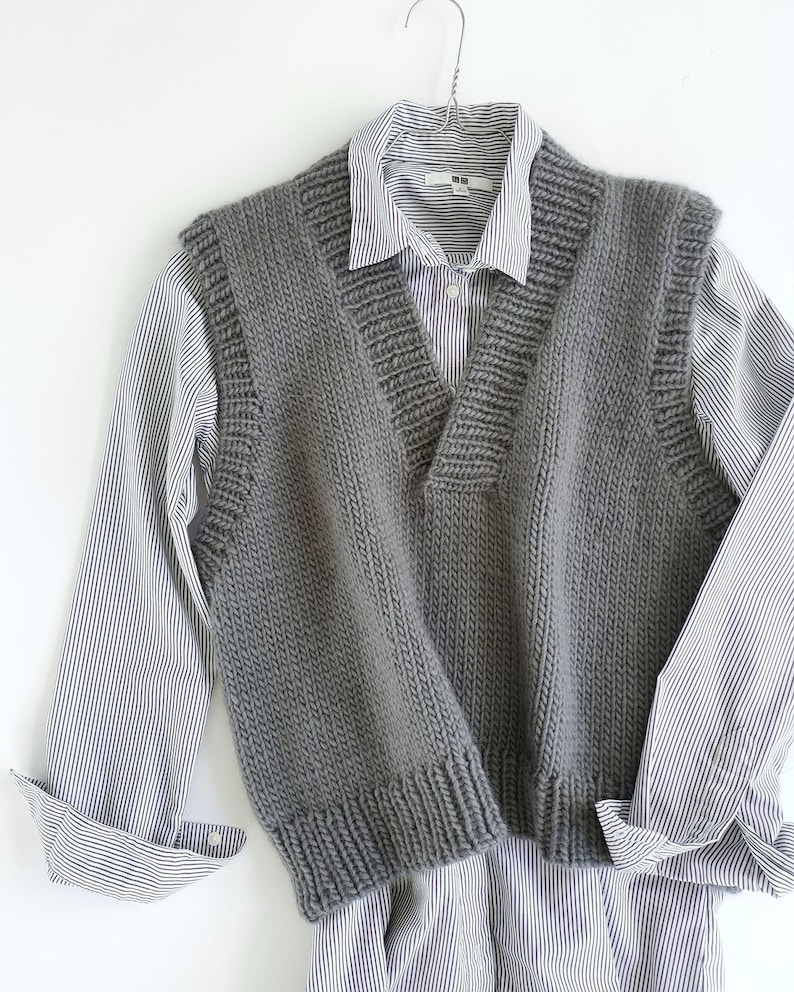 May include: A gray knitted vest with a V-neck and a ribbed texture. The vest is hanging on a hanger and is layered over a striped shirt.