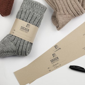 Pode incluir: Dois pares de meias de tric&ocirc; &agrave; m&atilde;o, cinzas e bege, com etiquetas de papel castanho. As etiquetas t&ecirc;m o texto "SOCKEN HANDGESTRICKT" e informa&ccedil;&otilde;es sobre a composi&ccedil;&atilde;o do material e as instru&ccedil;&otilde;es de cuidado. Uma etiqueta tem o texto "FOR" e "VON" preenchido com texto escrito &agrave; m&atilde;o.