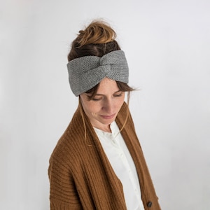 Könnte beinhalten: Ein graues Strick Stirnband mit einem verdrehten Detail. Das Stirnband wird von einer Person mit braunen Haaren und einem braunen Cardigan getragen.