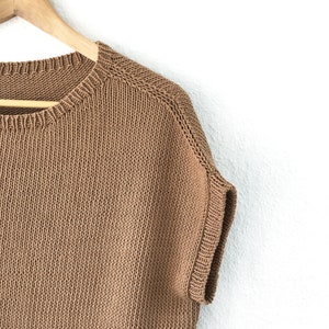 Pu&ograve; includere: Un maglione di lana marrone a maniche corte appeso a un appendiabiti di legno su uno sfondo bianco.