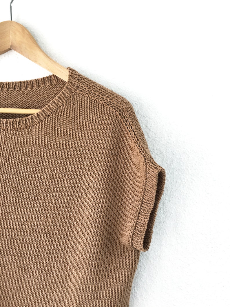 Pu&ograve; includere: Un maglione di lana marrone a maniche corte appeso a un appendiabiti di legno su uno sfondo bianco.