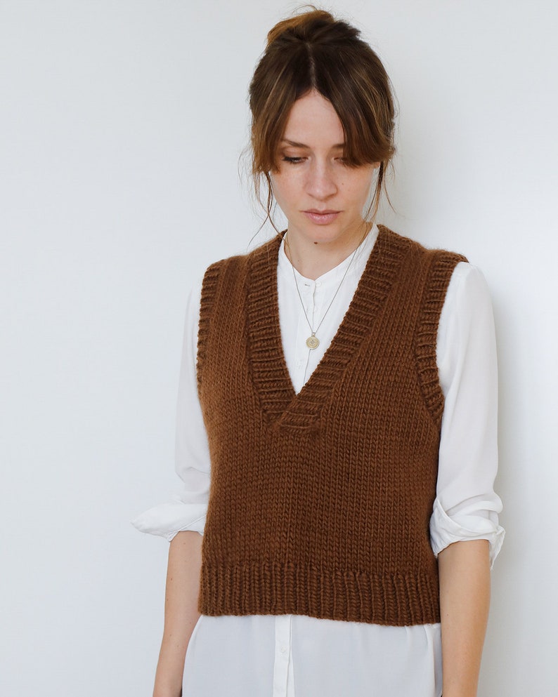 May include: A brown knitted vest with a V-neck and a ribbed hem. The vest is worn over a white button-down shirt.