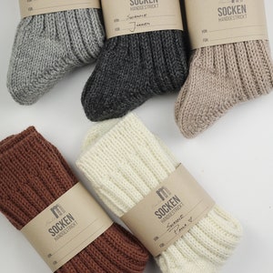 May include: Five pairs of hand-knitted socks in different colors, including gray, brown, beige, white, and cream. Each pair has a brown paper tag with the word "SOCKEN" and "HANDGESTRICKT" written on it. Some tags have handwritten names and a "FOR" section.