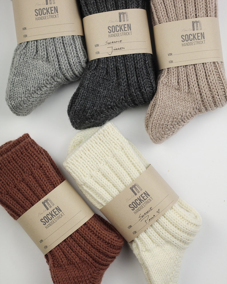 May include: Five pairs of hand-knitted socks in different colors, including gray, brown, beige, white, and cream. Each pair has a brown paper tag with the word "SOCKEN" and "HANDGESTRICKT" written on it. Some tags have handwritten names and a "FOR" section.