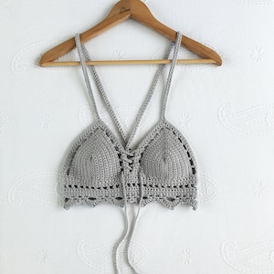 以下が含まれることがあります： 木製のハンガーにかけられた、グレーのクロシェのクロップトップ。前面にリボンがあり、リボンにはタッセルが付いています。