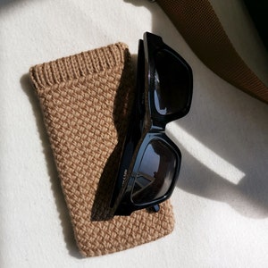 以下が含まれることがあります： 白い表面に、黒いサングラスがベージュのニットケースに入っています。サングラスは長方形のフレームです。背景には茶色のストラップが見えます。ケースは織り込まれた素材で作られています。