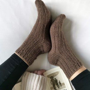 May include: A pair of brown knitted socks with a ribbed pattern. The socks are worn by a person sitting on a white surface with a book in their lap. The book cover features a black and white photo of a woman holding an umbrella.