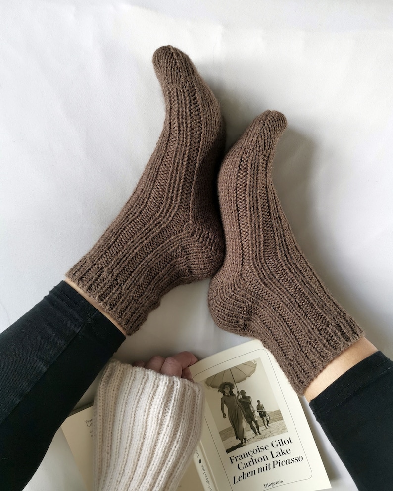 May include: A pair of brown knitted socks with a ribbed pattern. The socks are worn by a person sitting on a white surface with a book in their lap. The book cover features a black and white photo of a woman holding an umbrella.