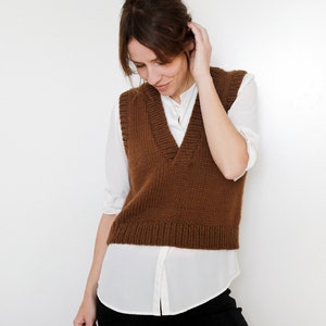 May include: A brown knitted vest with a V-neckline. The vest is worn over a white button-down shirt and black pants.