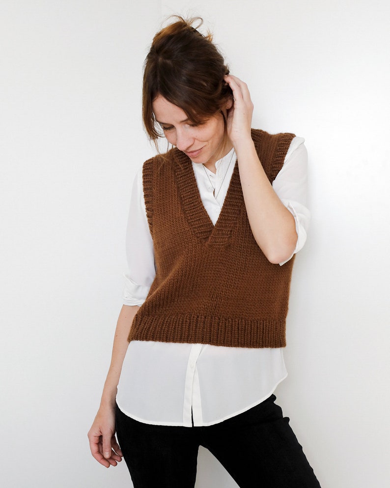 May include: A brown knitted vest with a V-neckline. The vest is worn over a white button-down shirt and black pants.