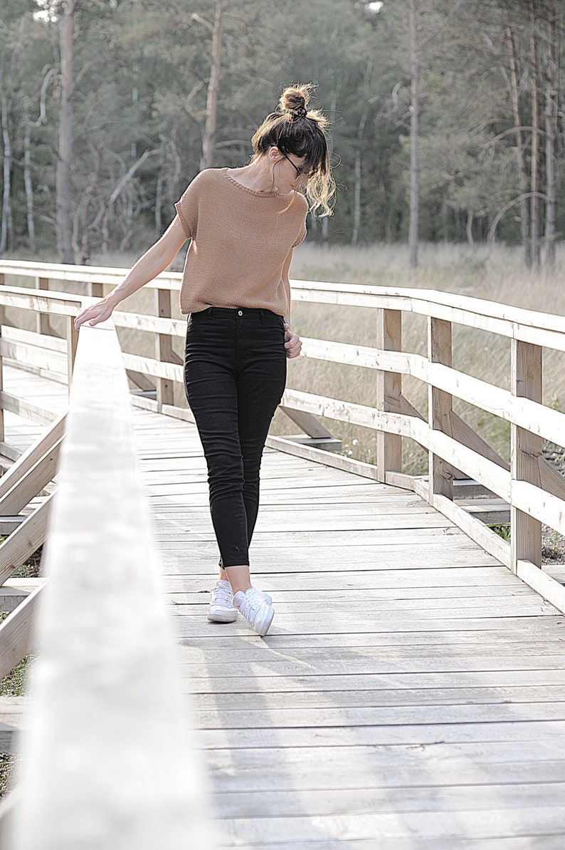 Pu&ograve; includere: Una donna con un maglione marrone e jeans neri cammina su un ponte di legno. Indossa scarpe da ginnastica bianche.