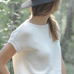 Pu&ograve; includere: Un top bianco a maniche corte in maglia con scollo rotondo. La modella indossa un cappello nero.