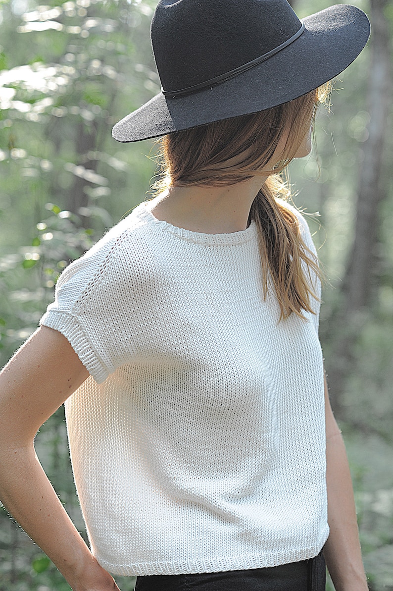 Pu&ograve; includere: Un top bianco a maniche corte in maglia con scollo rotondo. La modella indossa un cappello nero.