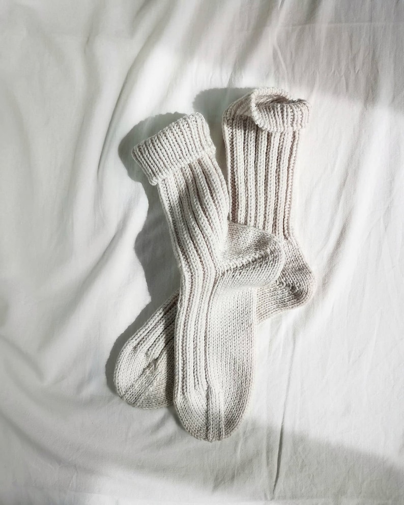 May include: A pair of off-white knitted socks with a ribbed texture. The socks are folded and laid out on a white fabric background.