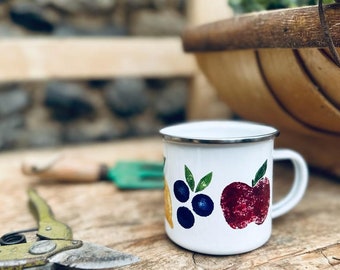 May include: White enamel mug with a red apple, yellow lemon, and blue blueberries painted on it. The mug has a silver rim and handle.