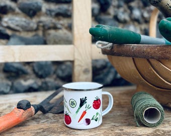 May include: White enamel mug with a silver rim, featuring illustrations of vegetables such as tomatoes, carrots, and radishes. The mug sits on a wooden surface, with gardening tools and a basket visible in the background.