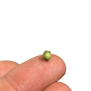 May include: A miniature light green frog figurine with a rounded body, small protruding limbs, and two prominent white eyes with black pupils. The tiny creature is sculpted with a smooth texture and sits upright, displaying a simple and compact form.