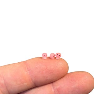 May include: Three small pink figurines with different facial expressions. The figurines are standing on a finger.